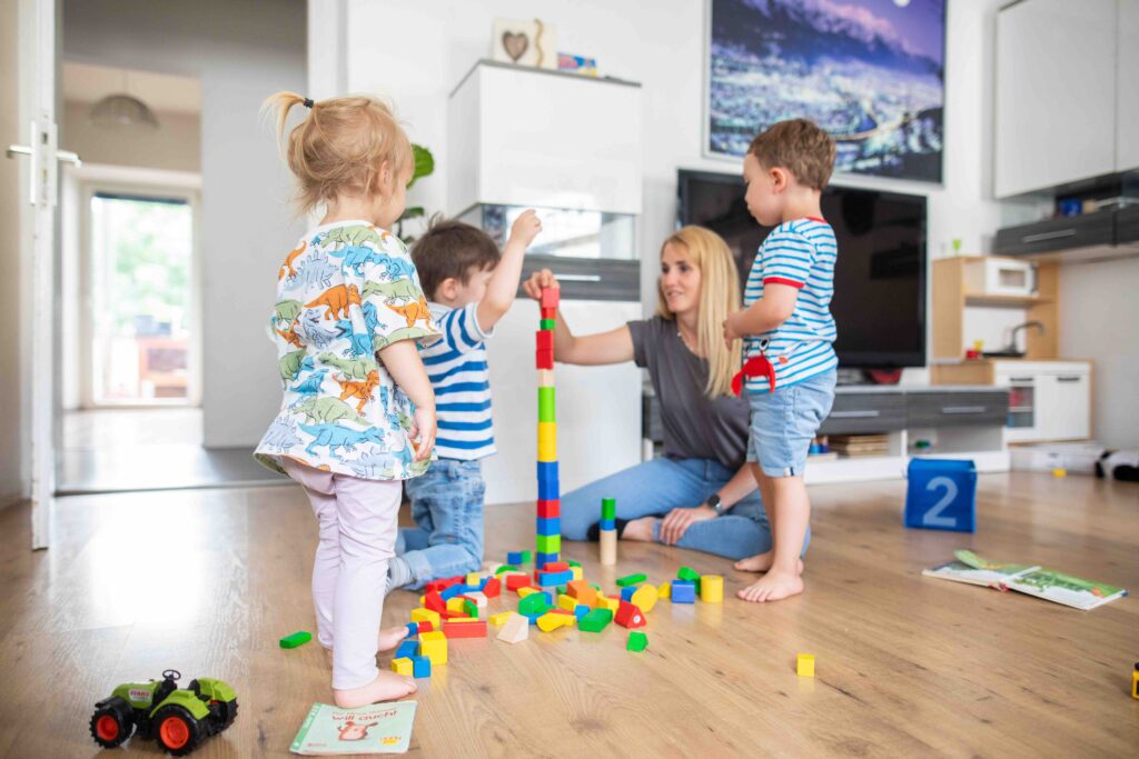 Eine Tagesmutter spielt mit Tageskindern im Wohnzimmer, sie spielen mit Bauklötzen.